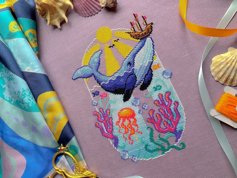 Font view of Whale with Boat cross stitch pattern. Blue whale has a boat on its nose that it has lifted out of the water. The sun is shining. Underwater we can see coral, fish, seaweed and a jellyfish. Colors are very vibrant. Birds are in the sky.
