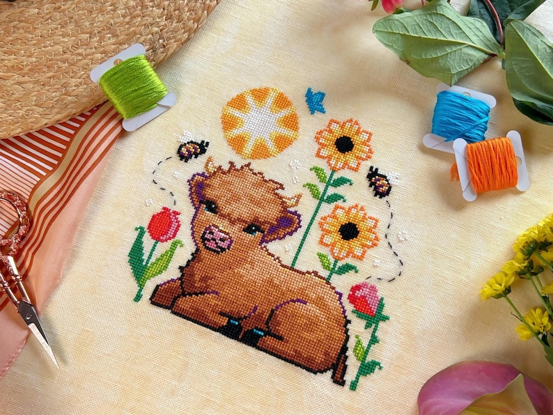 May include: Cross stitch pattern featuring a brown Highland cow calf with a yellow sun, sunflowers, and bees. The pattern is on a cream-colored fabric.
