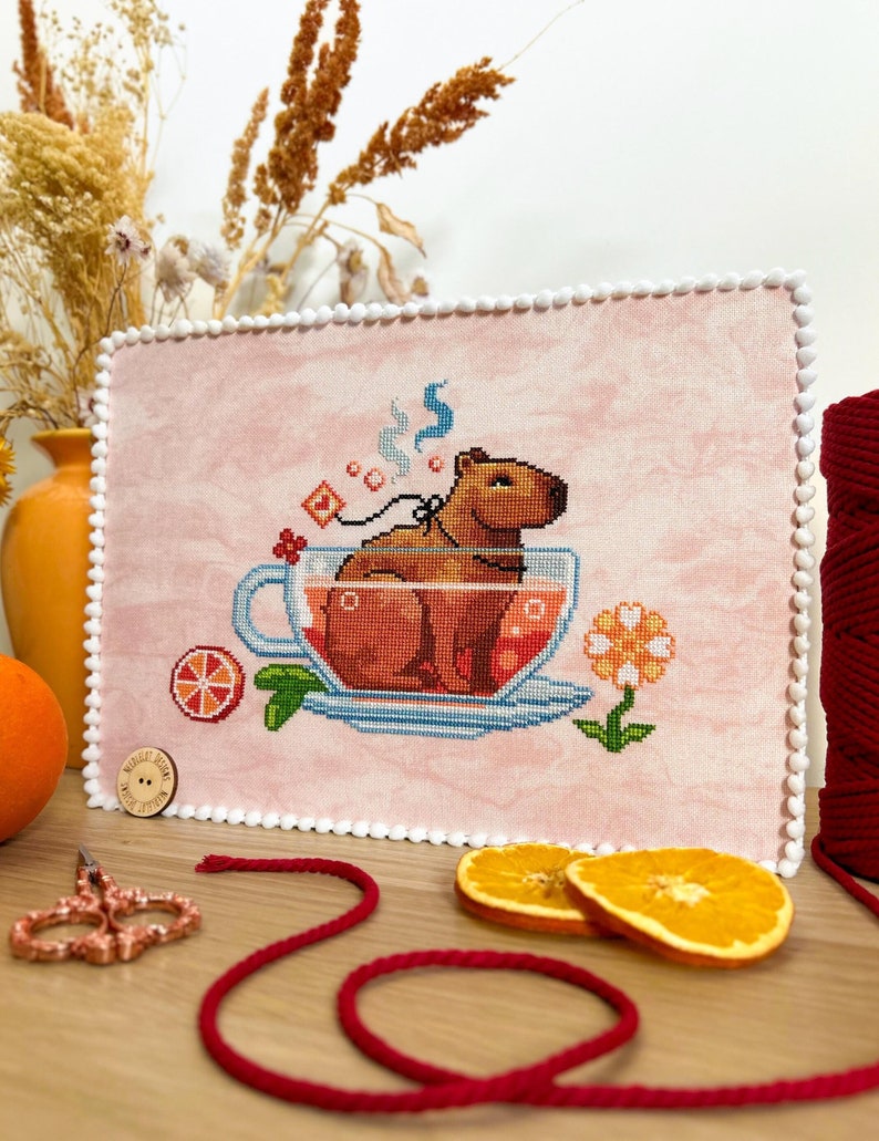K&ouml;nnte beinhalten: Ein Kreuzstichmuster mit einem Capybara, das in einer Teetasse mit Orangenscheiben und einer Blume sitzt. Der Hintergrund ist rosa und die Teetasse ist mit einer roten Fl&uuml;ssigkeit gef&uuml;llt. Das Muster ist mit einem wei&szlig;en Rahmen versehen.
