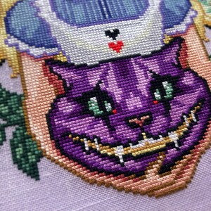 Closeup of Cheshire cat head that is placed in Alices arms. Stitches are neat and tidy. Cheshire cat is a vibrant purple with various different shades and markings in its face. Its nose is heart shaped. Its teeth are long and pointy. He looks cute.