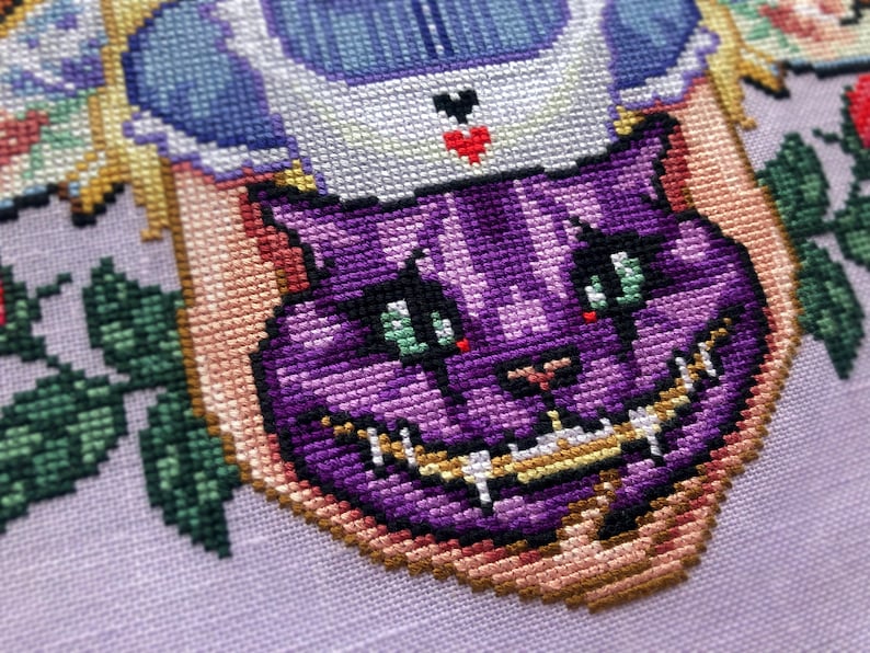 Closeup of Cheshire cat head that is placed in Alices arms. Stitches are neat and tidy. Cheshire cat is a vibrant purple with various different shades and markings in its face. Its nose is heart shaped. Its teeth are long and pointy. He looks cute.