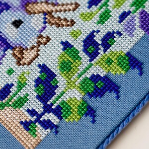 Puede incluir: Primer plano de un dise&ntilde;o de punto de cruz con un p&aacute;jaro y elementos florales. El bordado utiliza una paleta de azules, verdes y morados sobre un fondo azul claro. Un cord&oacute;n azul enmarca el borde de la tela.
