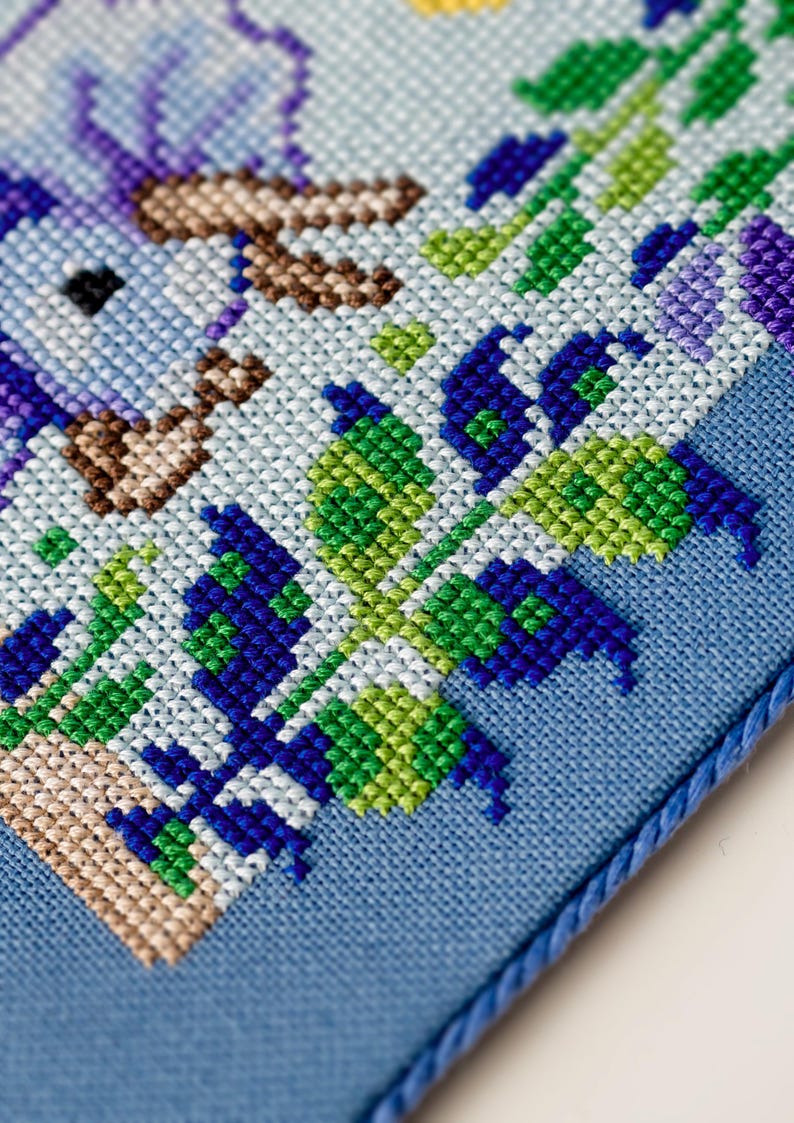 Puede incluir: Primer plano de un dise&ntilde;o de punto de cruz con un p&aacute;jaro y elementos florales. El bordado utiliza una paleta de azules, verdes y morados sobre un fondo azul claro. Un cord&oacute;n azul enmarca el borde de la tela.