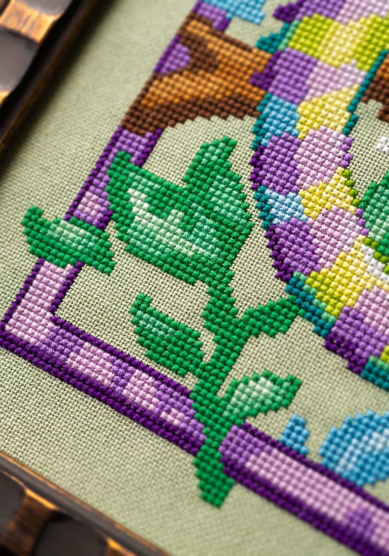 May include: Close-up of a cross-stitch design featuring a green leafy stem and purple border on a light green fabric. The design incorporates shades of green, purple, yellow, and blue, framed in a wooden border.