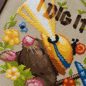 Peut inclure: Une broderie au point de croix sur tissu beige repr&eacute;sentant une taupe brune portant un casque de chantier jaune et tenant une pelle. Le texte "DIG IT" est brod&eacute; en brun au-dessus de la taupe, entour&eacute; de fleurs color&eacute;es, de feuilles vertes et d'un petit papillon bleu.