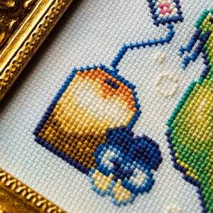 May include: A cross-stitch embroidery piece featuring a tea bag with an orange and yellow gradient, a blue tag, and a small blue flower, all set against a light blue fabric background and encased in a decorative, ornate gold-colored frame.