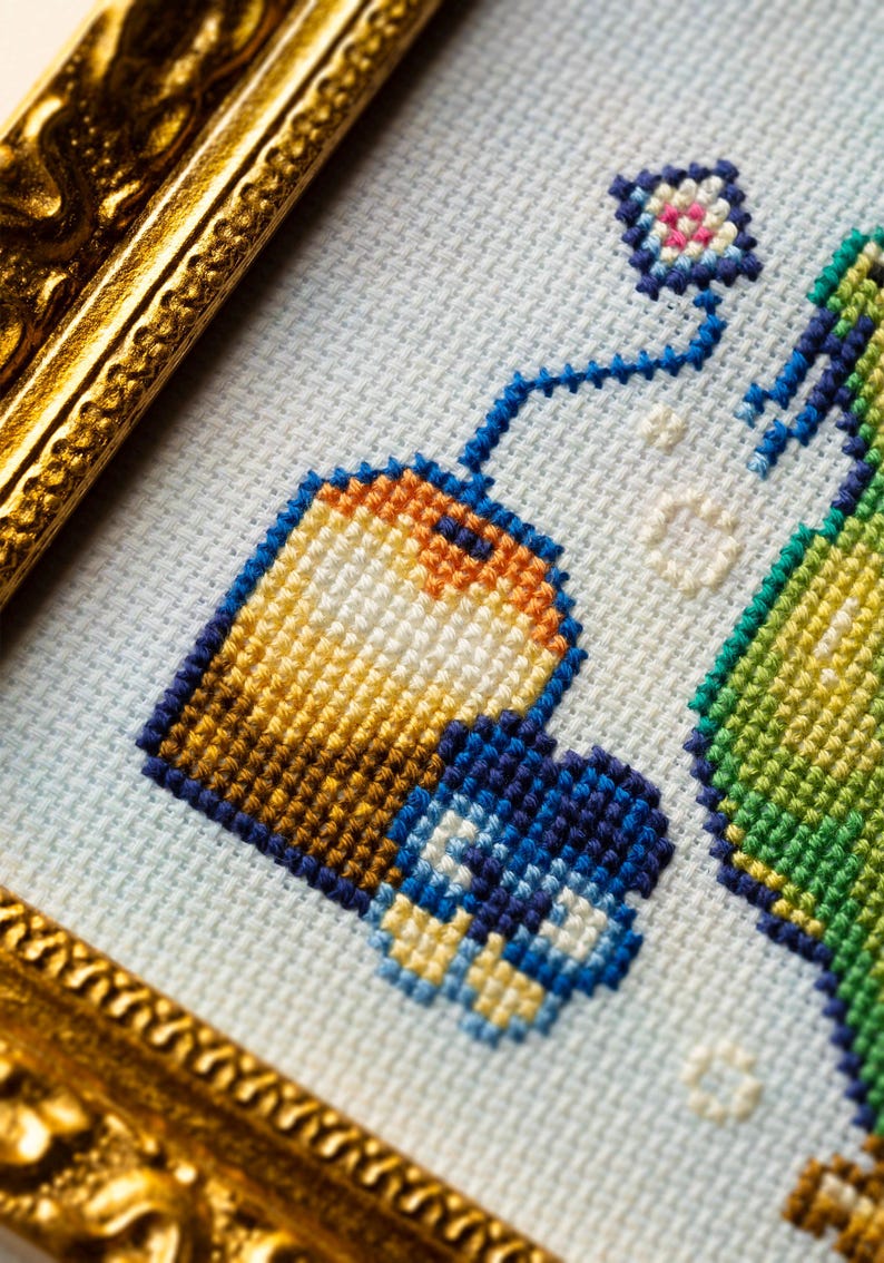 May include: A cross-stitch embroidery piece featuring a tea bag with an orange and yellow gradient, a blue tag, and a small blue flower, all set against a light blue fabric background and encased in a decorative, ornate gold-colored frame.