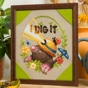 Peut inclure: Une broderie au point de croix encadr&eacute;e repr&eacute;sentant une taupe brune portant un casque de chantier jaune avec un faisceau lumineux blanc, entour&eacute;e de fleurs et de verdure. Le texte "I DIG IT" est brod&eacute; en lettres marron fonc&eacute;.