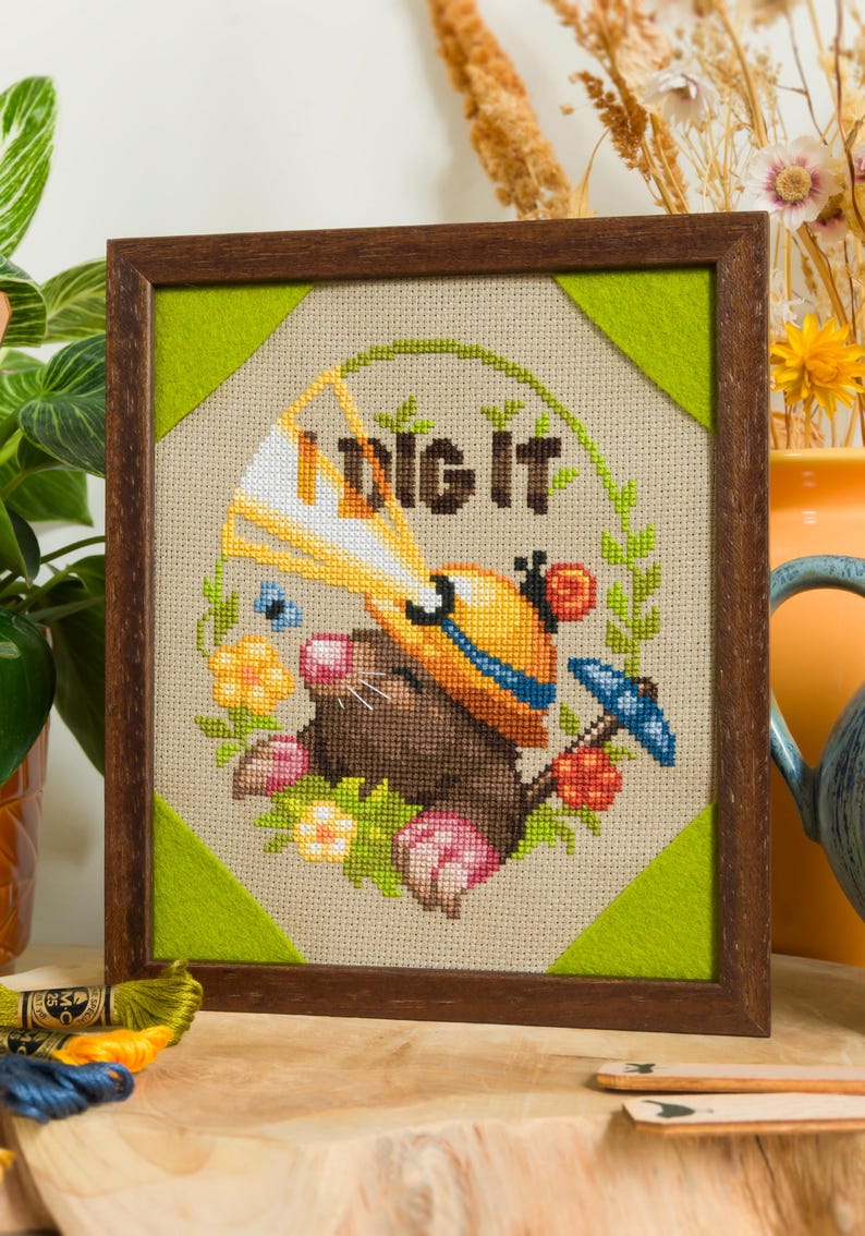Peut inclure: Une broderie au point de croix encadr&eacute;e repr&eacute;sentant une taupe brune portant un casque de chantier jaune avec un faisceau lumineux blanc, entour&eacute;e de fleurs et de verdure. Le texte "I DIG IT" est brod&eacute; en lettres marron fonc&eacute;.