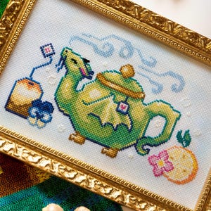 May include: A cross-stitch embroidery in a gold ornate frame depicting a green dragon-shaped teapot with steam, a tea bag, and a citrus slice with a pink flower. The design features vibrant green, yellow, orange, and blue thread colors on white fabric.