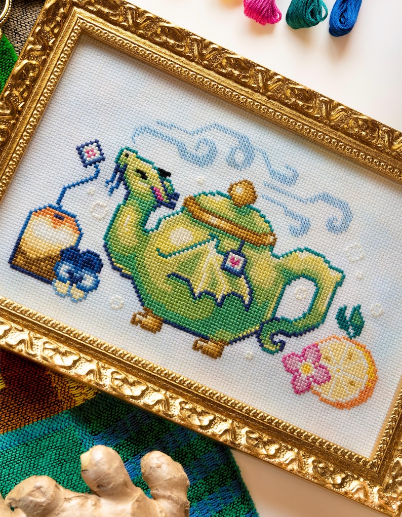 May include: A cross-stitch embroidery in a gold ornate frame depicting a green dragon-shaped teapot with steam, a tea bag, and a citrus slice with a pink flower. The design features vibrant green, yellow, orange, and blue thread colors on white fabric.