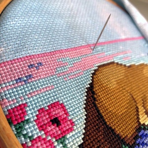 Work in progress on Platypus cross stitch pattern. Needle is visible and is stiching through the 16ct Aida fabric. The pink and blue sky with birds is being stitched. Stitches are fluffy and tidy. Danish stitching method is used.