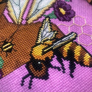 Closeup view of Queen Bee cross stitch pattern. Underneath the Queen Bee is an actual bee, of much smaller size. The bee is flying and is yellow with black brown stripes. Colors are vibrant and stitches are neat and tidy. Queens skin is a deep brown