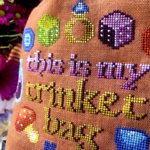Closeup of the Trinket Bag. Individual stitches are clearly visible. The colors are mainly brown, gold, purple, blue, red and yellow. Font reminds of adventure books or medieval times.