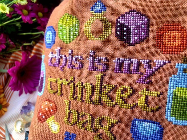 Closeup of the Trinket Bag. Individual stitches are clearly visible. The colors are mainly brown, gold, purple, blue, red and yellow. Font reminds of adventure books or medieval times.