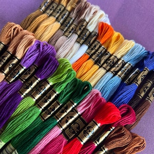 May include: A collection of DMC embroidery floss spools in various colors, including red, green, blue, purple, and brown. The spools are arranged in a fan shape, with the labels facing the viewer. The labels feature the DMC logo and the number of the floss color.