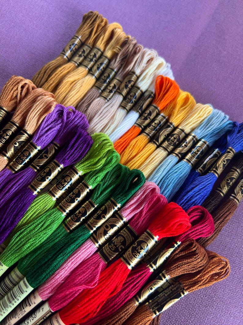 May include: A collection of DMC embroidery floss spools in various colors, including red, green, blue, purple, and brown. The spools are arranged in a fan shape, with the labels facing the viewer. The labels feature the DMC logo and the number of the floss color.