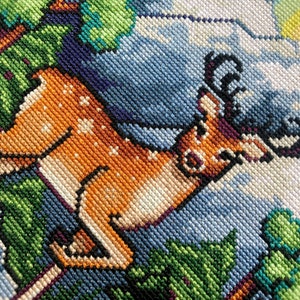 Puede incluir: Un primer plano de un bordado de punto de cruz que representa un ciervo marr&oacute;n con manchas blancas corriendo por un bosque verde. El ciervo tiene grandes cuernos y est&aacute; rodeado de follaje verde.