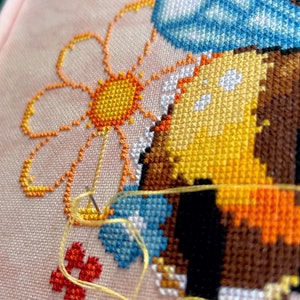 May include: Close-up of a cross-stitch embroidery project depicting a bee with blue wings and a yellow and orange body. The bee is near a yellow and orange daisy with a white center. The embroidery is on a pink fabric with a yellow thread.