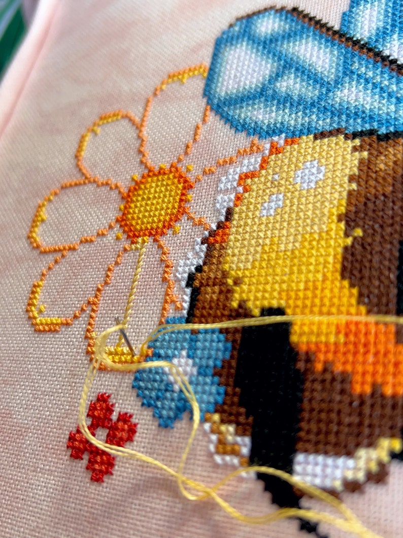 May include: Close-up of a cross-stitch embroidery project depicting a bee with blue wings and a yellow and orange body. The bee is near a yellow and orange daisy with a white center. The embroidery is on a pink fabric with a yellow thread.