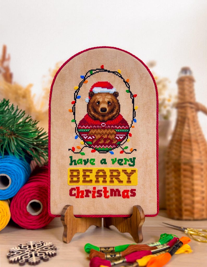 May include: A cross-stitch artwork featuring a brown bear wearing a Santa hat and a red sweater, surrounded by Christmas lights. The text reads "have a very BEARY Christmas" in green, yellow, and red. The artwork is mounted on a wooden stand.
