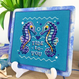 May include: Cross-stitch artwork featuring two seahorses facing each other, with the text "Tide - to - You" in white. The seahorses are purple, teal, and yellow on a turquoise background, framed in a marbled blue and white border. The artwork is displayed on a white stand.