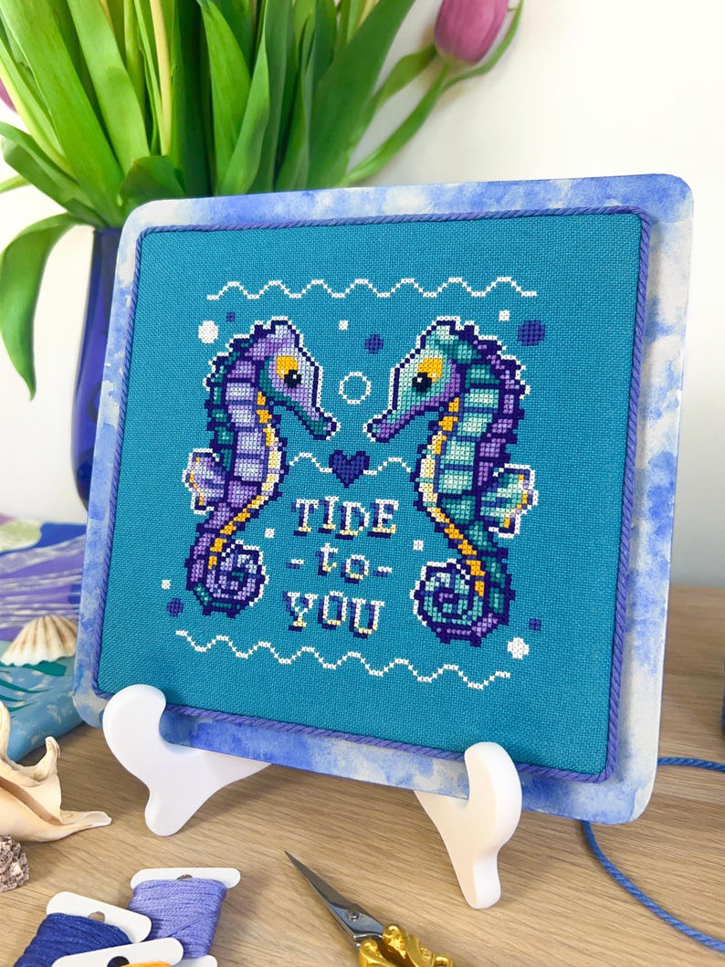 May include: Cross-stitch artwork featuring two seahorses facing each other, with the text "Tide - to - You" in white. The seahorses are purple, teal, and yellow on a turquoise background, framed in a marbled blue and white border. The artwork is displayed on a white stand.