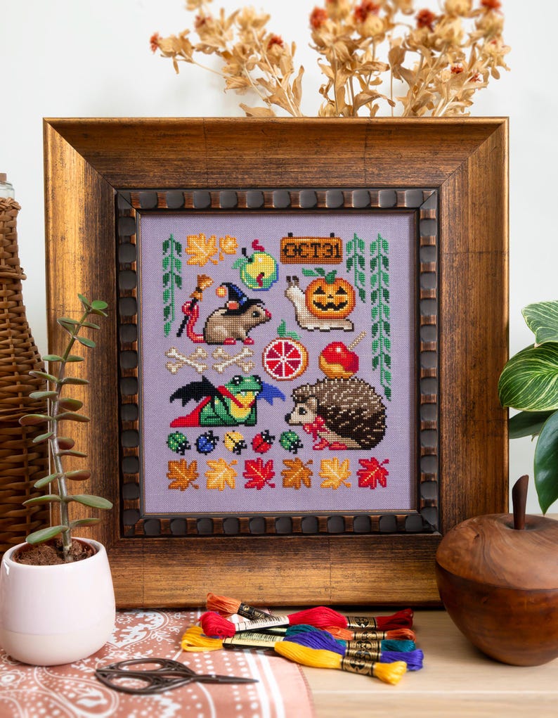 May include: Cross-stitch artwork in a wooden frame featuring Halloween-themed designs. The artwork includes a mouse in a witch hat, a pumpkin, a hedgehog, and various autumn leaves. The color palette includes purple, orange, green, and brown.