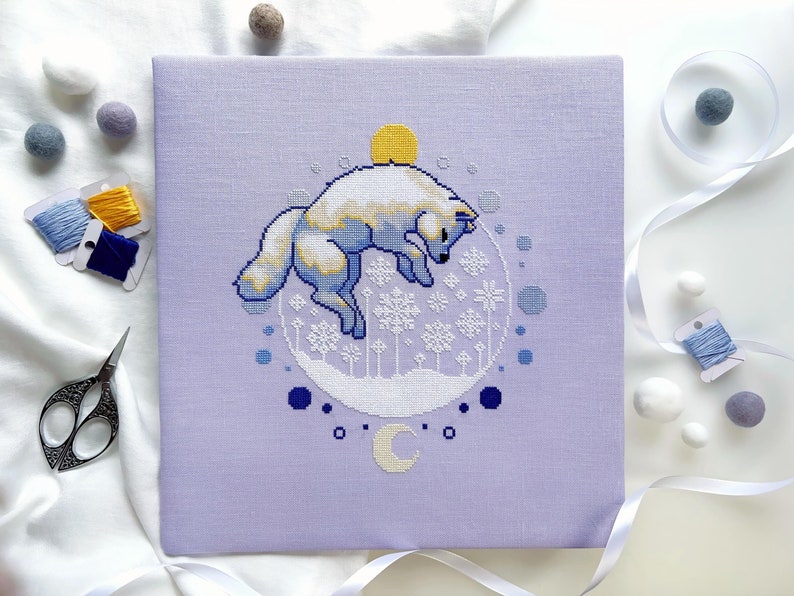 May include: A cross-stitch pattern on a purple fabric background featuring a white fox jumping in a circle of snowflakes. The fox is surrounded by a blue and white circle with a crescent moon and stars.