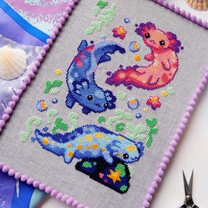 May include: Cross-stitch art featuring three axolotls in shades of blue, pink, and orange, with yellow and green accents. The artwork is framed with a lavender pom-pom trim. The background is a light gray fabric.