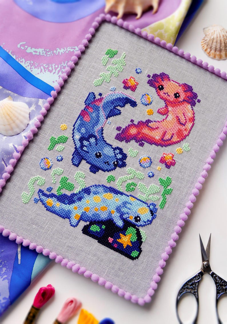 May include: Cross-stitch art featuring three axolotls in shades of blue, pink, and orange, with yellow and green accents. The artwork is framed with a lavender pom-pom trim. The background is a light gray fabric.