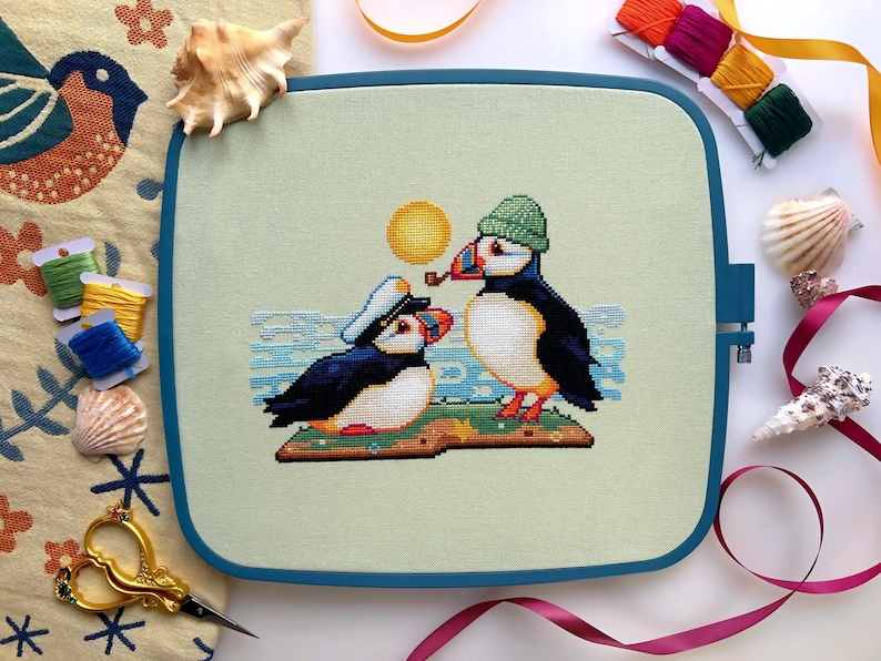 May include: Cross-stitch embroidery of two puffins on a green background. The puffin on the left is wearing a blue sailor hat and the puffin on the right is wearing a green hat. The puffin on the right is holding a pipe in its beak.