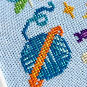 May include: Close-up of a cross-stitch design on light blue fabric. The design features a blue and orange hot air balloon, green and blue dragonfly, and yellow stars. The needlework is detailed and colorful.