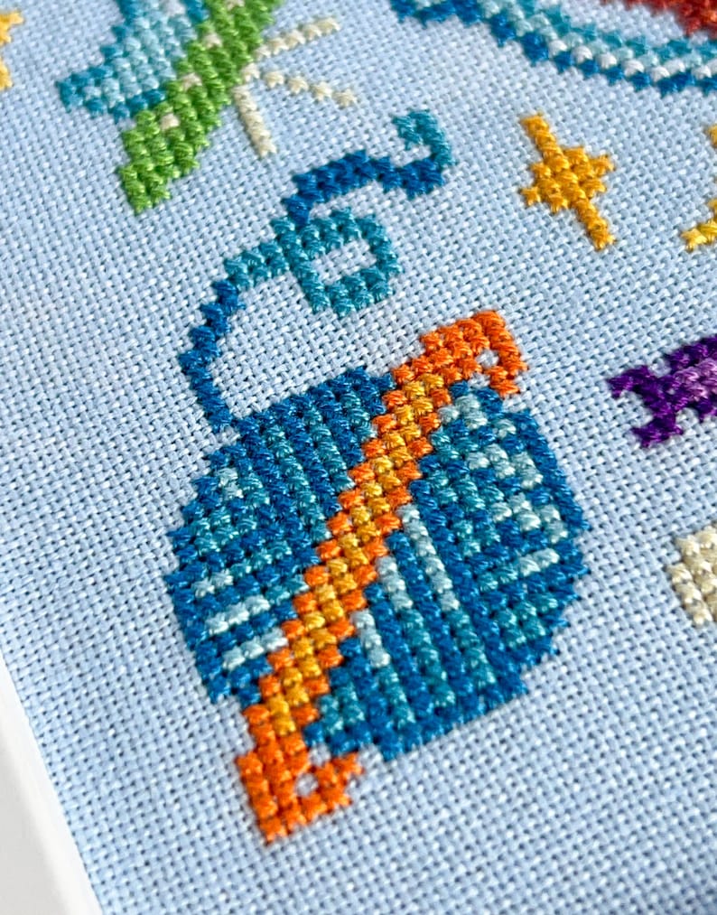 May include: Close-up of a cross-stitch design on light blue fabric. The design features a blue and orange hot air balloon, green and blue dragonfly, and yellow stars. The needlework is detailed and colorful.