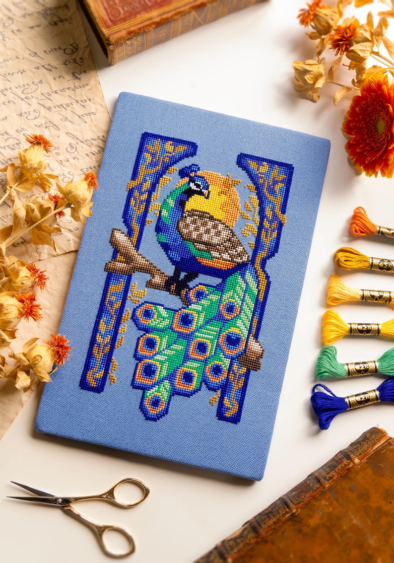 May include: A rectangular blue fabric panel featuring a detailed cross-stitch embroidery of a peacock perched on a branch. The peacock displays vibrant blue, green, and yellow plumage. The design is framed by a stylized golden yellow border.