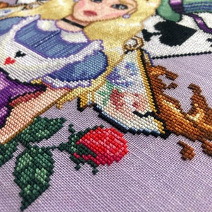 Closeup of righthand bottom side of Alice in Wonderland cross stitch pattern. One of the roses and teacups are particularly visible here. The teacup is a traditional English teacup, which brown tea is spilling out of. The rose is red and pink.