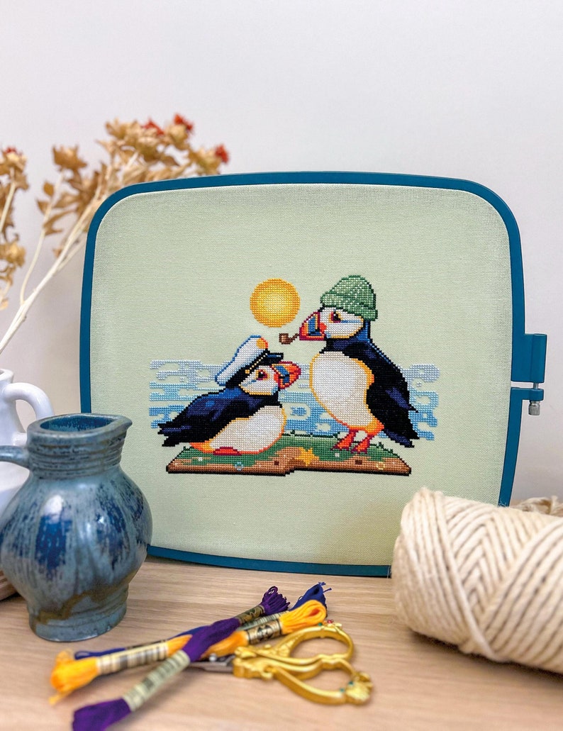 May include: Cross-stitch embroidery of two puffins wearing hats, one with a pipe, on a green background with a yellow sun and blue waves. The puffins are sitting on a book.