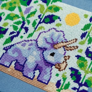 Puede incluir: Dise&ntilde;o de punto de cruz con un triceratops morado con cuernos marrones, rodeado de follaje verde y morado, y un sol amarillo sobre un fondo azul claro. El dise&ntilde;o est&aacute; sobre una tela azul.