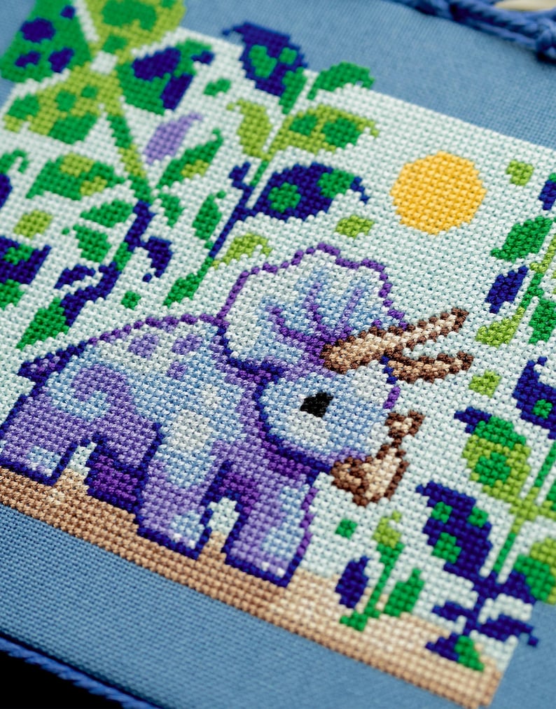 Puede incluir: Dise&ntilde;o de punto de cruz con un triceratops morado con cuernos marrones, rodeado de follaje verde y morado, y un sol amarillo sobre un fondo azul claro. El dise&ntilde;o est&aacute; sobre una tela azul.