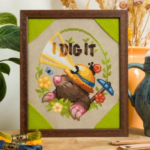 Peut inclure: Une broderie au point de croix encadr&eacute;e repr&eacute;sentant une taupe brune portant un casque de chantier jaune et tenant une pelle jaune. Le texte "I DIG IT" est brod&eacute; au-dessus, entour&eacute; d'une couronne de feuilles vertes, de fleurs et d'un parapluie bleu.