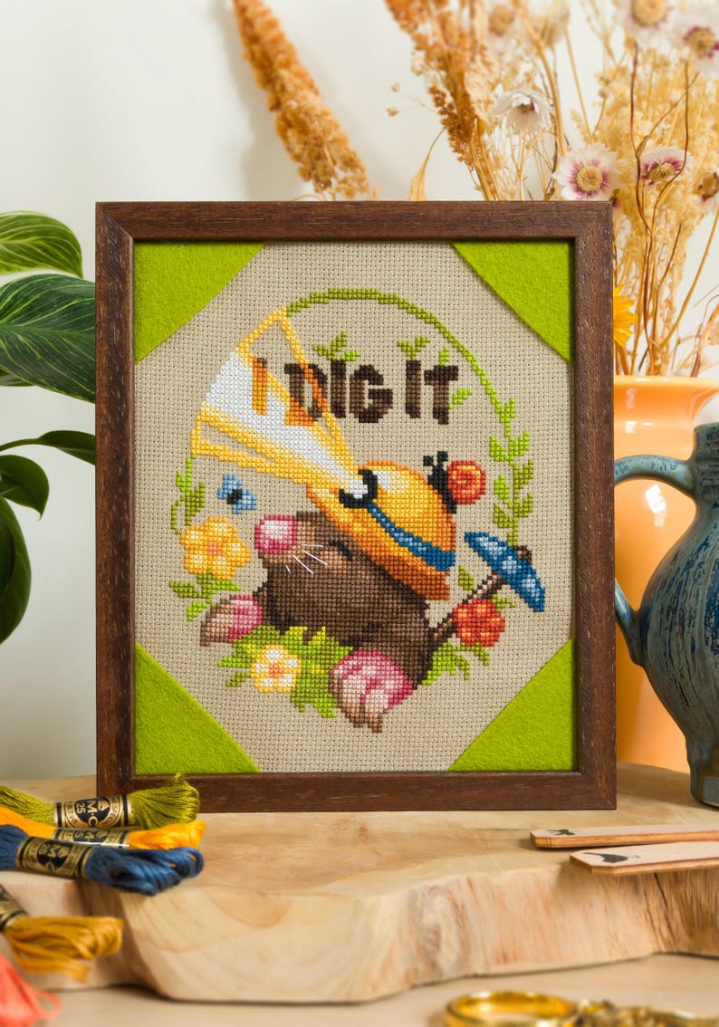Peut inclure: Une broderie au point de croix encadr&eacute;e repr&eacute;sentant une taupe brune portant un casque de chantier jaune et tenant une pelle jaune. Le texte "I DIG IT" est brod&eacute; au-dessus, entour&eacute; d'une couronne de feuilles vertes, de fleurs et d'un parapluie bleu.