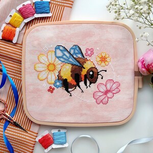 May include: A pink embroidery hoop with a cross-stitch design of a bumblebee surrounded by flowers. The bumblebee is yellow and black with blue wings. The flowers are pink, yellow, and white.