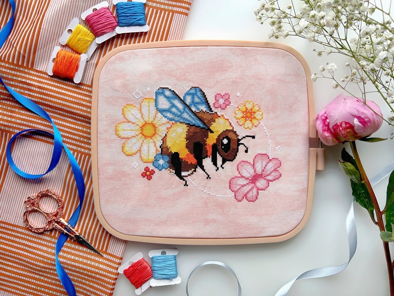 May include: A pink embroidery hoop with a cross-stitch design of a bumblebee surrounded by flowers. The bumblebee is yellow and black with blue wings. The flowers are pink, yellow, and white.