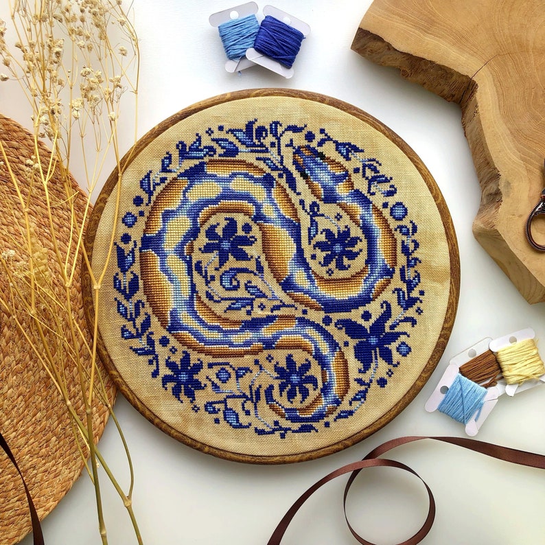Peut inclure: Un cercle de broderie circulaire avec un motif floral bleu et or repr&eacute;sentant un serpent stylis&eacute;. Le motif est sur un fond de tissu beige.