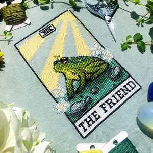 Flatlay of The Friend frog tarot cross stitch pattern. Stitched item, surrounded by decorations. Finished piece is of small to medium size. Colors are yellow, green, blue, grey and black. Frog is cute and small. The Friend is written underneath.