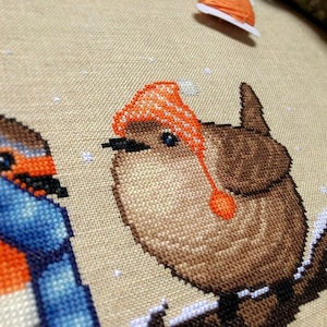 Closeup of Winter Birds cross stitch pattern. Image is zoomed in on the wren. He is very cute and round. The wren is wearing an orange, striped hat with pompoms, and looks very warm. The colors of his feathers are brown and white.