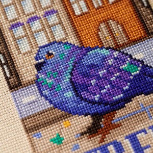 Puede incluir: Arte de punto de cruz que representa una paloma con plumaje azul y morado, posada frente a edificios. Los edificios est&aacute;n representados en tonos marrones y naranjas, con la palabra "STREET" visible.
