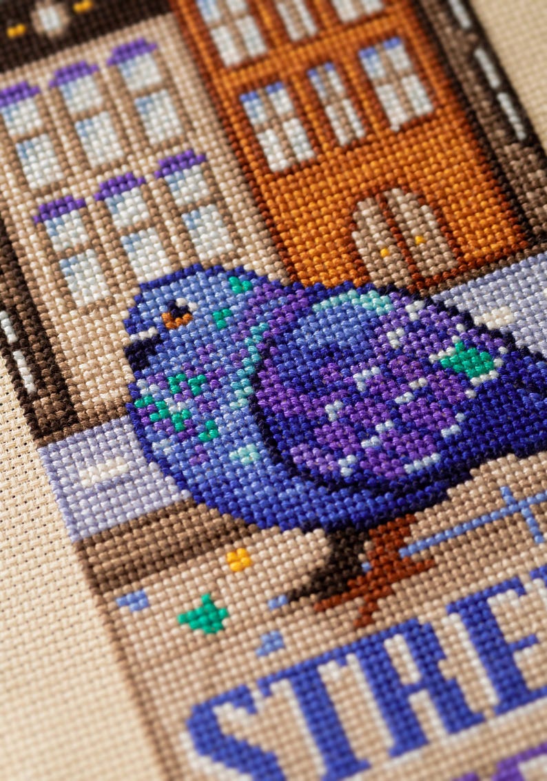 Puede incluir: Arte de punto de cruz que representa una paloma con plumaje azul y morado, posada frente a edificios. Los edificios est&aacute;n representados en tonos marrones y naranjas, con la palabra "STREET" visible.