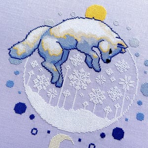 May include: Cross-stitch pattern featuring a white fox with blue accents jumping over a crescent moon and a yellow sun. The fox is surrounded by a circle of blue and white snowflakes.