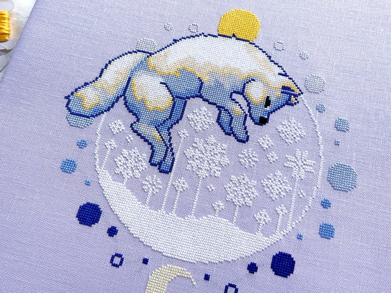 May include: Cross-stitch pattern featuring a white fox with blue accents jumping over a crescent moon and a yellow sun. The fox is surrounded by a circle of blue and white snowflakes.